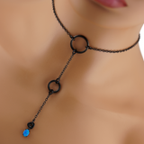 A close-up image of a Blue Opal BDSM O-Ring Lariat Necklace featuring a lab-created blue opal pendant, seamless metal O-rings, and a black stainless steel rolo chain, displayed on a mannequin.